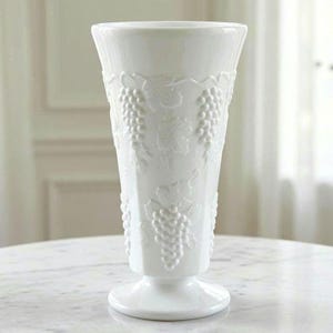 May include: A tall, white milk glass vase featuring a flared rim and a pedestal base. The exterior surface is decorated with a raised, embossed pattern of grape clusters and vine leaves that wrap around the body of the vessel in a repeating design.