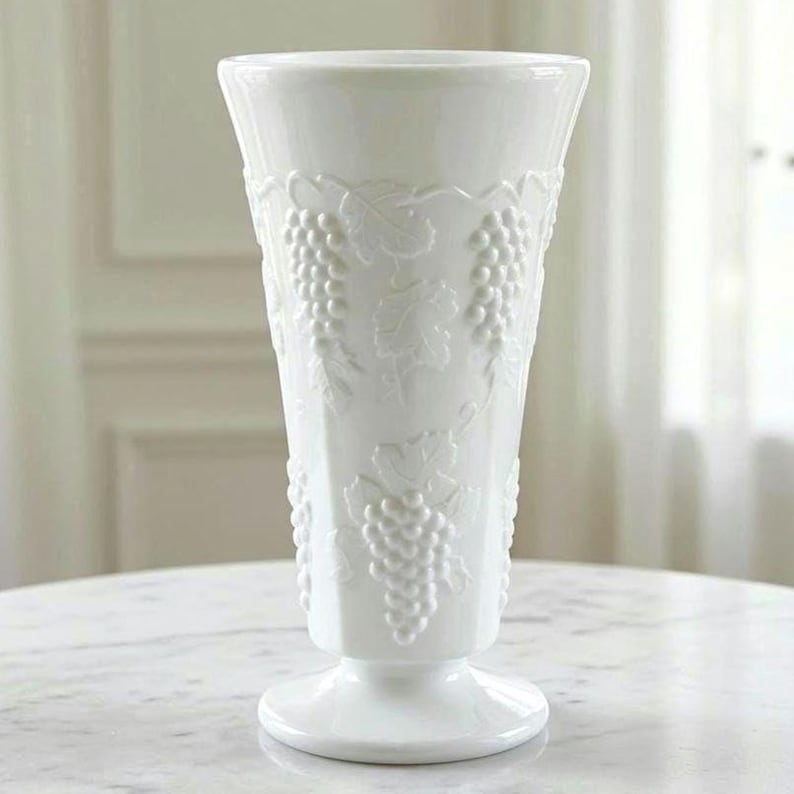May include: A tall, white milk glass vase featuring a flared rim and a pedestal base. The exterior surface is decorated with a raised, embossed pattern of grape clusters and vine leaves that wrap around the body of the vessel in a repeating design.
