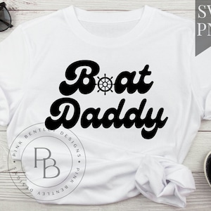 May include: White t-shirt with black text that reads "Boat Daddy" in a retro font. The word "Boat" has a small anchor symbol in the center of the letter "O".