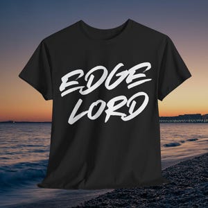 Op de afbeelding: Zwart t-shirt met de witte tekst "EDGE LORD" in een gedurfde, handgeschreven stijl. Het shirt wordt gepresenteerd tegen een achtergrond van een strand bij zonsondergang.