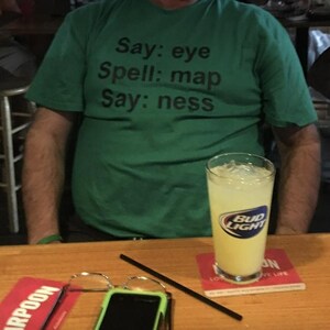 Puede incluir: Una persona lleva una camiseta verde con el texto "Say: eye Spell: map Say: ness" impreso en ella. La persona sostiene un vaso de limonada con un logotipo de Bud Light.