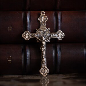 May include: A bronze-toned metal crucifix featuring an ornate cross with trefoil-shaped ends and a central figure of Jesus. The cross includes the letters INRI inscribed above the figure and intricate floral detailing at the center intersection.