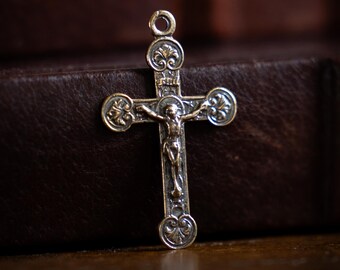May include: A silver-toned metal crucifix pendant featuring a detailed figure of Jesus on the cross. The cross has rounded ends decorated with embossed floral patterns and a small circular loop at the top for attachment to a chain or cord.
