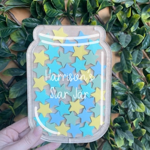 May include: A clear glass jar with a wooden lid filled with blue and yellow star-shaped pieces. The jar has the text "Harrison's Star Jar" written on it.