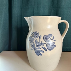 May include: Cream-colored ceramic pitcher with a blue floral design. The pitcher has a handle and a spout for pouring. The floral pattern includes stylized flowers and leaves. The pitcher is likely vintage or retro in style.