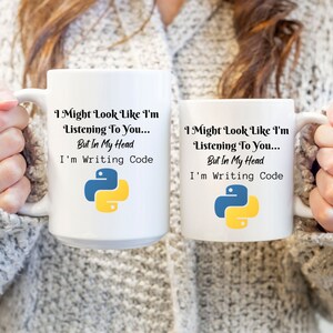 K&ouml;nnte beinhalten: Zwei wei&szlig;e Keramikbecher mit dem Text "I Might Look Like I'm Listening To You... But In My Head I'm Writing Code" und einem blau-gelben Python-Logo. Die Becher werden von einer Person gehalten, die einen grauen Pullover tr&auml;gt.