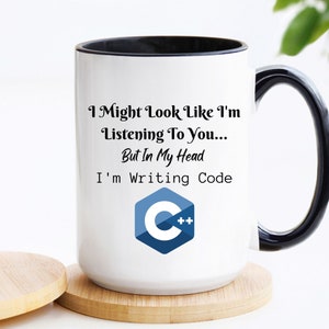 May include: White ceramic mug with a black rim and handle. The mug features a black and white graphic with the text "I Might Look Like I'm Listening To You... But In My Head I'm Writing Code" and a blue and white C++ logo.