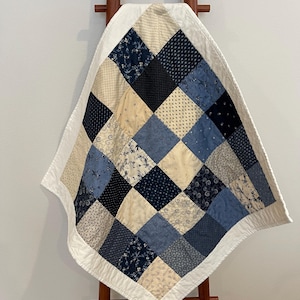 May include: A patchwork quilt with a white border, featuring squares in various shades of blue, cream, and navy. The quilt has a diamond pattern and is draped over a wooden ladder. The quilt's dimensions are approximately 40 inches by 40 inches.