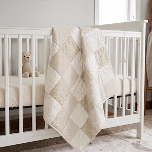May include: A patchwork quilt featuring a repeating diamond pattern in alternating shades of beige, cream, and white. The fabric squares display various subtle floral and geometric prints, creating a textured appearance draped over a white crib.