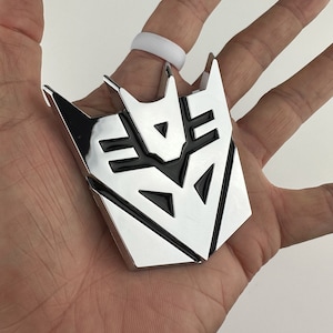 May include: A silver and black metal emblem with a geometric design. The emblem is shaped like a stylized robot head with a black triangular eye in the center.