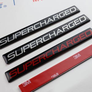 Puede incluir: Cuatro emblemas rectangulares negros con la palabra "SUPERCHARGED" en letras blancas y rojas. Los emblemas tienen un acabado brillante y están dispuestos en fila. Se ve una tira adhesiva roja 3M.