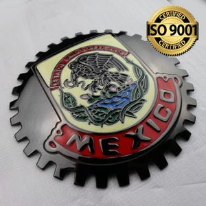 Może przedstawiać: Czarny metalowy emblemat samochodowy z złotym wzorem koła zębatego. Środek zawiera żółtą tarczę z czarnym orłem, zielonymi liśćmi i niebieską wodą. Tekst "ESTADOS UNIDOS MEXICANOS" jest napisany wokół tarczy. Tekst "MEXICO" jest napisany na czerwono wokół tarczy.