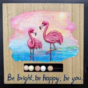 May include: A wooden wall art piece with a painted sunset scene featuring two pink flamingos. The flamingos are standing in a blue and green water scene. The art piece has a row of colorful beads at the bottom and the text "Be bright, be happy, be you."