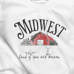 以下が含まれることがあります： 赤い納屋のグラフィックと「Midwest」の文字がプリントされた白いスウェットシャツ。「land of hopes and dreams」のテキストが下にあります。柔らかい素材で作られたスウェットシャツで、肌寒い日に最適です。