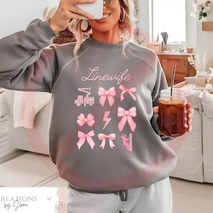 May include: Gray sweatshirt with the text "Linewife" in pink lettering. The sweatshirt features a design of pink bows, a lightning bolt, and a utility truck.