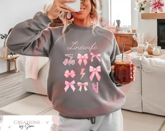 May include: Gray sweatshirt with the text "Linewife" in pink lettering. The sweatshirt features a design of pink bows, a lightning bolt, and a utility truck.