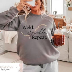 May include: A gray sweatshirt with the text "Weld. Sparkle. Repeat." in black script and a gold chain design. The person is holding a glass jar with a drink and a straw. The sweatshirt is part of a clothing collection.