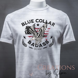 May include: White t-shirt featuring a graphic design with the text "BLUE COLLAR BADASS" above an eagle illustration, with an American flag background. The text "DIRTY HANDS. STILL BROKE." is below the eagle. The shirt is displayed on a mannequin.