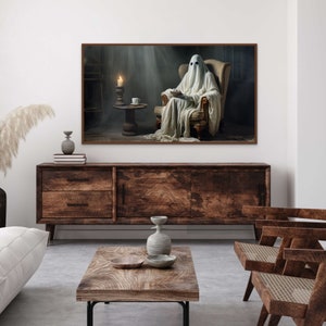 May include: A white ghost figure sits in a brown armchair, reading a book. A lit candle and a cup sit on a small table in the background. The image is framed in a brown wood frame.