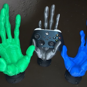 Könnte beinhalten: Drei 3D-gedruckte Hände, eine grüne, eine silberne und eine blaue, halten einen schwarzen Xbox-Controller.