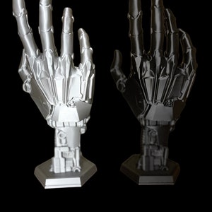 Peut inclure: Deux mains robotiques imprimées en 3D, l'une argentée et l'autre noire, sont posées avec les doigts tendus. Les mains sont montées sur des bases hexagonales.