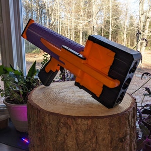 May include: A purple and orange toy gun with a black magazine. The gun is sitting on a wooden stump.