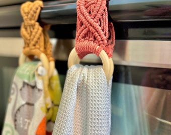 Pode incluir: Dois panos de cozinha pendurados na pega de um forno preto. Um tem um topo em macramé amarelo mostarda com estampado de gato, o outro um topo em macramé rosa seco com uma toalha de tecido cinzento claro texturizado anexada.