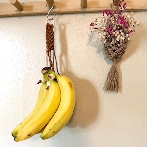 May include: A bunch of bananas hanging from a macrame banana holder with white beads. The holder is made of brown cord and is hanging from a hook on a wooden wall.