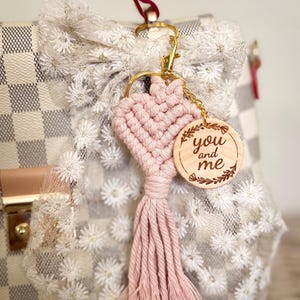 Puede incluir: Un llavero de corazón de macramé rosa con una etiqueta de madera que dice "you and me". El llavero está sujeto a un cierre dorado y a un bolso a cuadros blanco y beige.