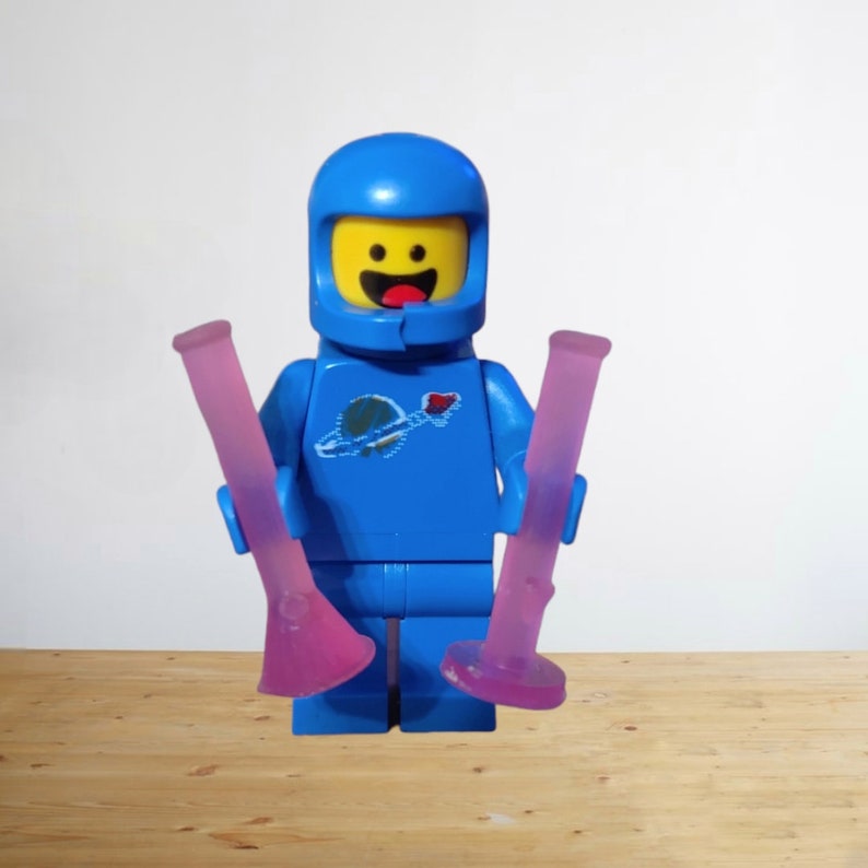 May include: A blue Lego astronaut figure with a yellow face and a smiling expression. The astronaut is holding two pink glass bongs. The astronaut has a planet and rocket ship logo on the chest.