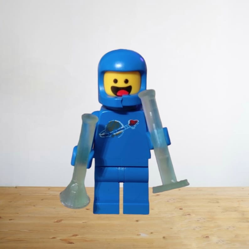 May include: A blue Lego astronaut figure with a yellow face and a blue helmet, holding two translucent, light blue glass tubes. The astronaut has a planet and rocket ship emblem on the chest.