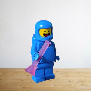 May include: A blue Lego astronaut figure with a yellow face and a pink, elongated object. The astronaut is wearing a blue helmet and suit. The background is a plain white wall and a wooden surface.