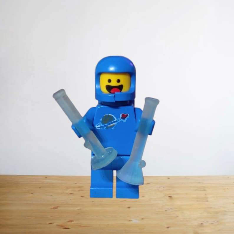 May include: A blue Lego astronaut figure with a yellow face and a blue helmet, holding two clear glass bongs. The astronaut has a logo on the chest. The bongs are clear with blue accents.