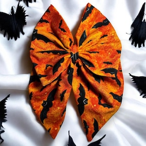 May include: A large orange and black Halloween hair bow with a bat pattern. The bow is surrounded by black bat decorations on a white background. The bow is made of fabric and has a star and moon pattern.