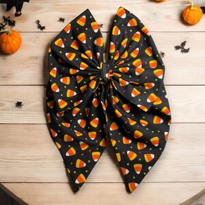 May include: A black bow with a candy corn pattern. The bow is made of fabric and has a pointed shape.
