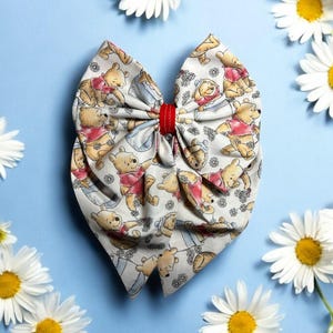 May include: A white fabric bow with a red ribbon in the center. The bow is decorated with a pattern of Winnie the Pooh characters. The bow is surrounded by white daisies on a light blue background.