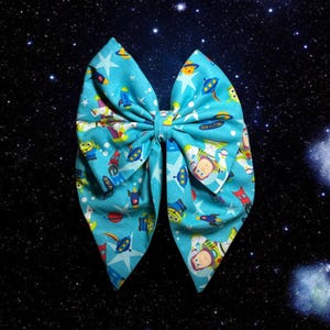 May include: A blue bow with a space-themed print featuring characters from the Toy Story franchise. The bow is against a black background with white stars.