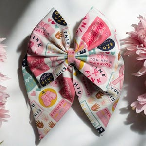 May include: A pink and white fabric bow with a pink ribbon in the center. The fabric is printed with various pink and white designs and text, including "Think Pink", "You Are A Warrior", and "Give Up Never".