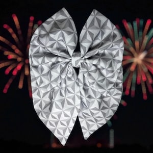 Könnte beinhalten: Eine große, grau-weiß gemusterte Schleife vor einem dunklen Hintergrund mit farbenfrohem Feuerwerk. Die Schleife hat ein geometrisches Design. Das Feuerwerk zeigt rote, orange und grüne Explosionen und schafft eine festliche Atmosphäre.