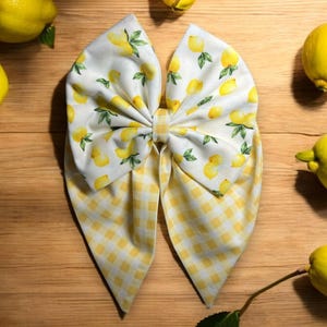 May include: A yellow and white bow with a lemon print. The bow is made of two pieces of fabric, one with a checkered pattern and the other with a lemon print. The bow is tied in a knot in the center.