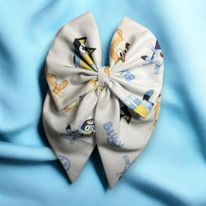 May include: A large, light gray fabric bow featuring cartoon characters in blue, yellow, and brown. The bow is tied in the center and has the word "Bluey" printed on it. The bow is set against a light blue fabric background.