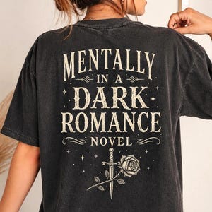 May include: Dark gray t-shirt with the text "MENTALLY IN A DARK ROMANCE NOVEL" in a vintage font. The design includes a dagger, rose, and stars. The shirt has a slightly faded, worn look.