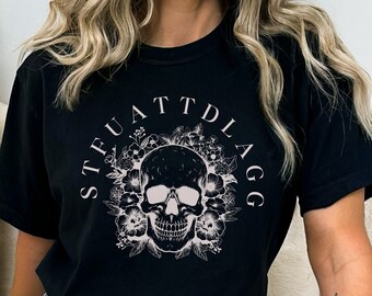 May include: Black t-shirt featuring a white skull and floral design. The text "STFUATT DLAGG" is written in a circle around the skull.