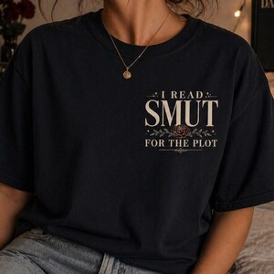 May include: Black t-shirt with the text "I READ SMUT FOR THE PLOT" in a decorative font. The shirt has a crew neck and short sleeves. The text is in a serif font with a floral design.