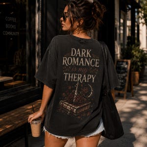 Könnte beinhalten: Ein anthrazitfarbenes Oversize-T-Shirt mit einem Grafikdruck auf der Rückseite, der den Text "DARK ROMANCE is my THERAPY" über einer Abbildung eines geschlossenen Buches, eines Dolches, roter Rosen und einer Mondsichel zeigt. Es hat kurze Ärmel.