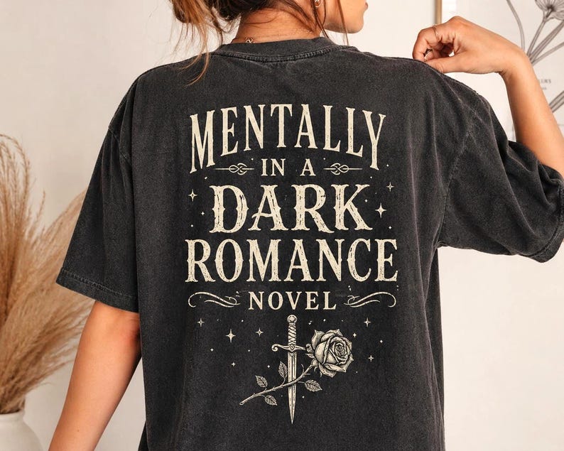 May include: Dark gray t-shirt with the text "MENTALLY IN A DARK ROMANCE NOVEL" in a vintage font. The design includes a dagger, rose, and stars. The shirt has a slightly faded, worn look.