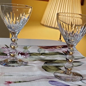May include: Two clear glass wine glasses with a cut glass design and a beaded stem. The glasses are sitting on a white tablecloth with a floral pattern.