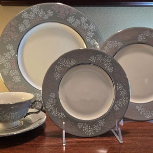 May include: A set of four gray and white floral patterned ceramic dinnerware pieces, including a large dinner plate, a smaller dinner plate, a salad plate, and a teacup and saucer.