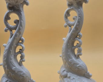 May include: Pair of white ceramic candle holders with a dolphin-like design. The candle holders have a gold trim on the base and around the top. The design features ornate curves and details, creating an elegant aesthetic.