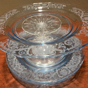 May include: A stack of light blue glass plates with a decorative white floral pattern around the edges. A clear glass cake stand is in the center. The plates are stacked on a brown surface.
